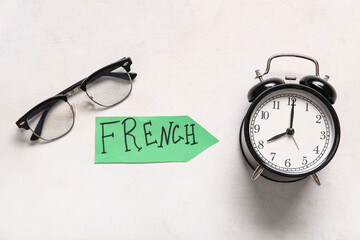 Paper sheet with word FRENCH, eyeglasses and alarm clock on light background