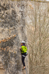 A woman is climbing a rock wall in a green jacket. She is wearing a helmet and a black backpack