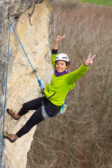 A woman is rock climbing and is wearing a green jacket. She is smiling and has her hands up in the air