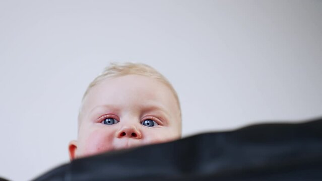 Lovely blond baby peeping out from behind the sofa. Serious grey eyed infant kid portrait. Low angle view.