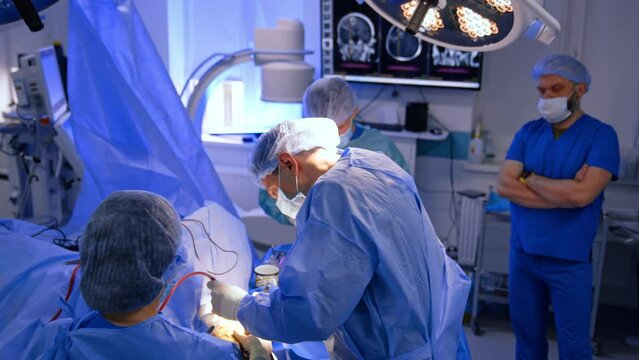Professional male neurosurgeon carrying out the operation assisted by two medics. Anesthesiologist stands at backdrop looking at screen with MRI scans.