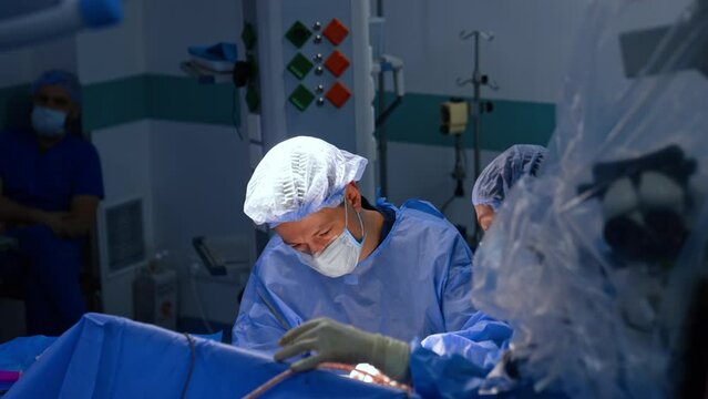 Two doctors working at neurosurgery using metal tools. Male anesthesiologist sits at backdrop. Top view.