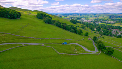 AERIAL: Tractor with trailer climbs a steeper road up a hill past green pastures