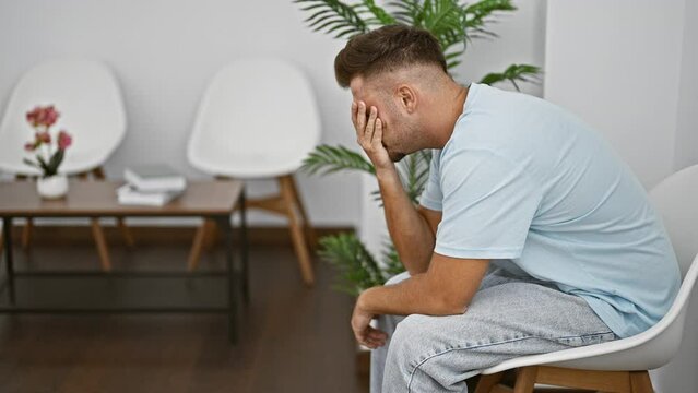 A distressed young hispanic man sitting in a clean modern indoor waiting room featuring white chairs and plants.
