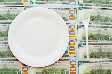 A disposable paper plate and a disposable fork on a background of hundred-dollar bills, business...