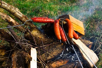 Traditional-style roasted sausages over a grassy bonfire with wood embers, emitting white smoke. Sausages exhibit a beautiful golden-brown color.