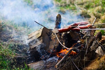 Traditional-style roasted sausages over a grassy bonfire with wood embers, emitting white smoke. Sausages exhibit a beautiful golden-brown color.