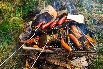 Traditional-style roasted sausages over a grassy bonfire with wood embers, emitting white smoke. Sausages exhibit a beautiful golden-brown color.