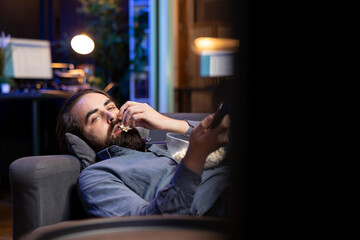 Bored supporter watching live football match broadcast, getting ready to change channel. Man laying down on couch holding remote control, eating popcorn and looking at soccer tournament