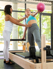 Fototapeta na wymiar Professional Latin female instructor helping girl practicing pilates exercising with small pilates ball at gym. Individual coach