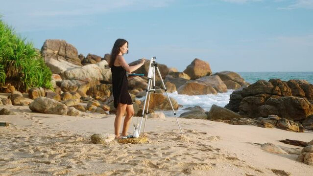 Artistic woman paints seascape on beach easel. Creative painter in black dress works on canvas against ocean backdrop. Art hobby, wave splashes. Inspiring coastal scene, rocky shore, blue sky. Slowmo