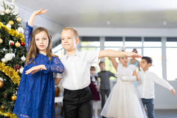 Smiling junior schoolchildren in party dresses learning waltz with pedagogue in hall embellished...