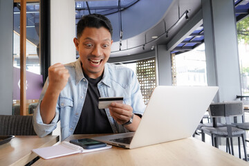 Asian man using laptop and mobile phone for shopping online with credit card in coffee shop online...