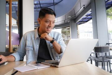 Asian man happy and smile face with blue shirt using laptop and mobile phone in coffee shop, online...