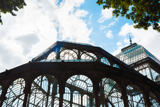 An intricate iron and glass canopy of a historic glasshouse looms against the cloud-dappled sky in a peaceful corner of Madrid