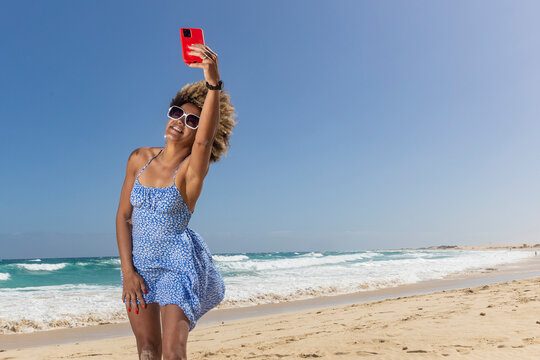 A woman enjoys capturing a breezy selfie by the ocean waves, their joy evident on a perfect, sunny beach day