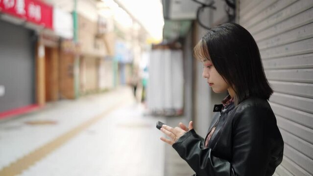 A young Japanese woman from Okinawa Prefecture in her 20s in winter clothes standing in a shopping street near Kokusai-dori Street in Naha City, Okinawa Prefecture and operating her smartphone 沖縄県那覇市の