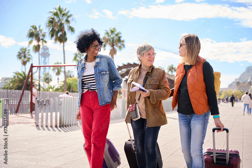 Wall mural Group mature diverse tourist women walking with their luggage while talking and looking at each other. Tourism of middle-aged people enjoying their journeys on the sunny city street. Copy space