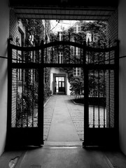 Black and white image of an open wrought iron gate leading to a courtyard with buildings, evoking a sense of welcome and mystery.