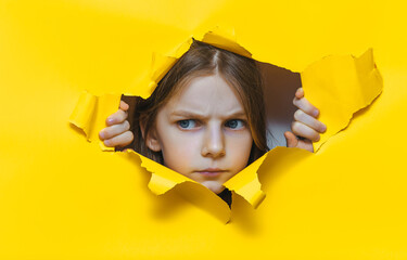 A funny and stern red-haired little girl looks disapprovingly through a hole in yellow paper. The concept of surprise, fear, fright, severe mood from what he saw. Anger and surprise. Copy space.