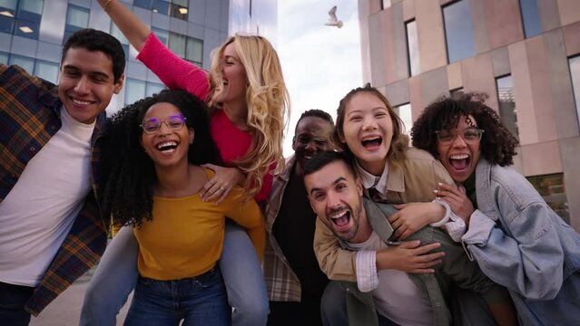 A group of multi-ethnic young friends are having fun in the city, sharing smiles and laughter. People doing piggyback cheerful looking at camera outdoor together. Friendship and community concept