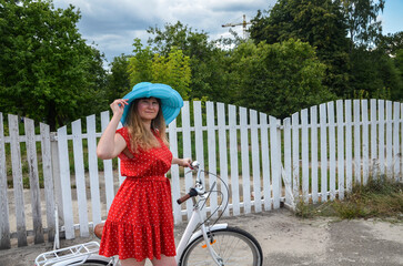 Young carefree woman in bright red dress and broad brim hat standing with bicycle and smiling