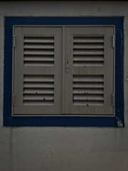 Closed gray shutters on a blue-framed window set in a textured dark wall, with a minimalist and...
