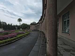 Obraz premium Pathway leading to a classical building with columns, surrounded by manicured gardens and lush greenery under a cloudy sky.