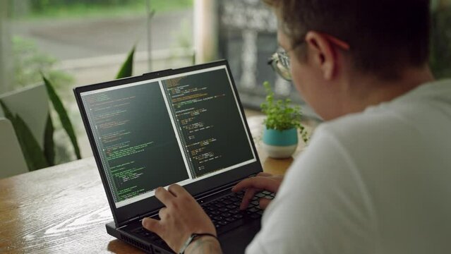 Transgender developer programs on laptop at plant-filled workspace. Codes, reviews scripts in casual coworking hub. Inclusive tech environment, diverse pro focused on computer tasks. Slow motion.