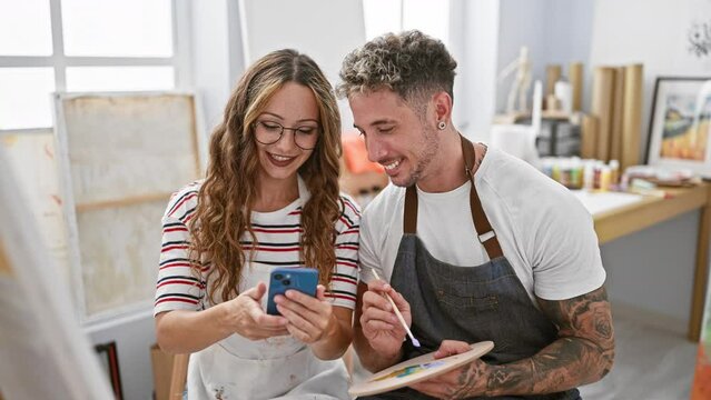 A woman and man, artists, enjoy a moment smiling with a phone selfie indoors at an art studio.