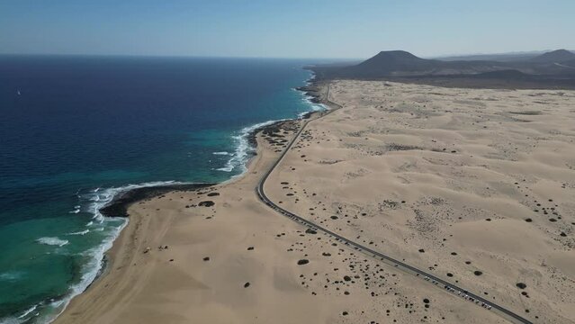 Aerial view of a single winding road cutting through the expansive Corralejo Dunes with the azure ocean gently lapping the shore in Fuerteventura