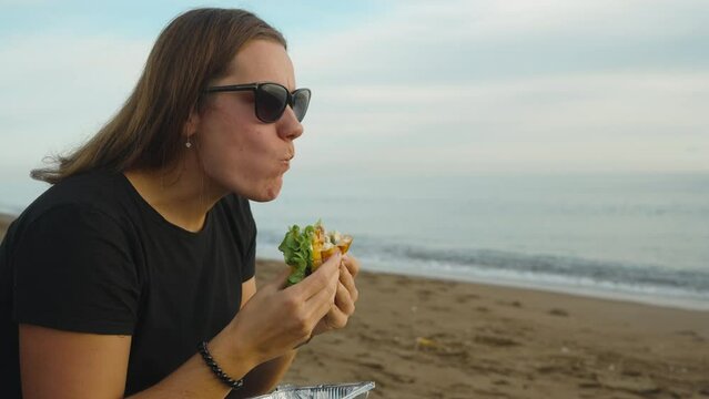 A young woman on the seashore, with her mouth wide open, bites off a large piece of burger. Picnic by the sea.