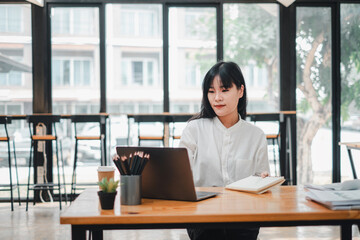 Young professional woman with a contemplative expression works at her laptop in a well lit cafe, surrounded by work materials.