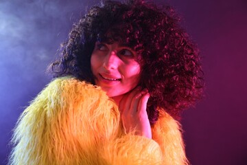 Fototapeta premium Beautiful young woman in yellow fur coat on color background in neon lights and smoke