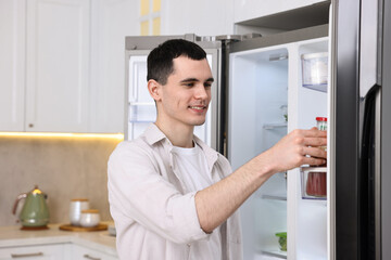 Naklejka na ściany i meble Happy man taking sauce out of refrigerator in kitchen