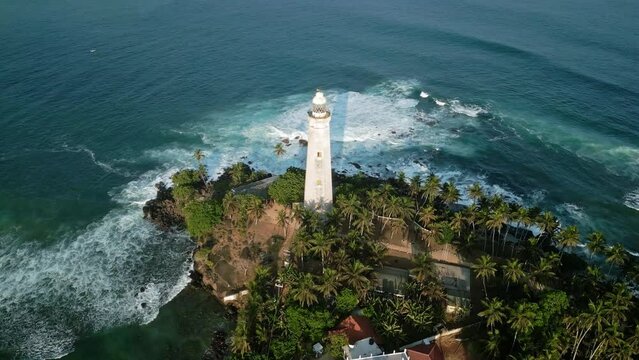 Drone circles historic Dondra lighthouse in Sri Lanka, surrounded by wild turquoise sea. Waves crash against rocky coast, greenery fringes structure, travel destination for history, nature lovers.
