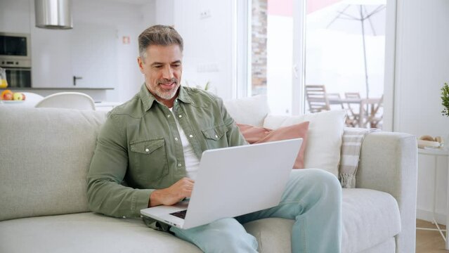 Happy middle aged man using laptop sitting on sofa at home. Mature older user looking at computer browsing internet, doing ecommerce shopping on website relaxing on couch in modern living room.