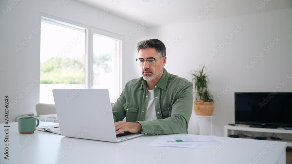 Wall mural Older mature middle aged man wearing eyeglasses looking at computer technology sitting at table, using laptop hybrid working online, elearning, browsing web, searching online at home. - Wall murals