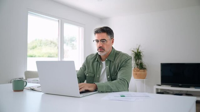 Older mature middle aged man wearing eyeglasses looking at computer technology sitting at table, using laptop hybrid working online, elearning, browsing web, searching online at home.