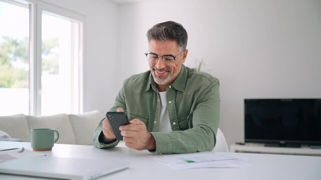 Smiling senior mature middle aged man wearing eyeglasses holding cell phone using smartphone sitting at home table, scrolling social media, checking financial apps, buying online, texting messages.
