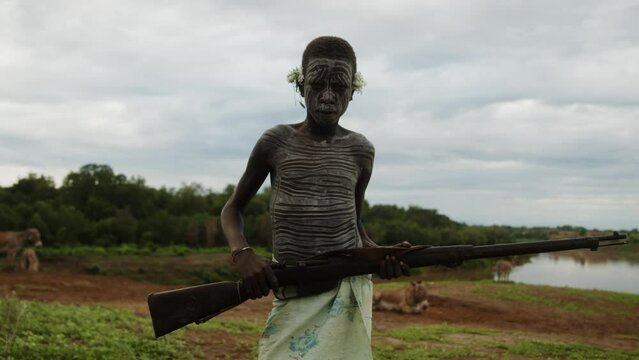 A Kid with Face Paint from Karo Tribe with a flower on his ear holding a rifle