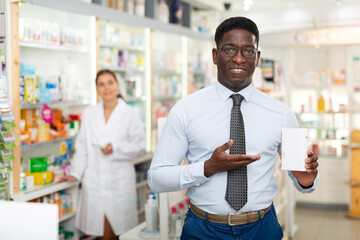 Fototapeta na wymiar African-american man standing in salseroom of drugstore and holding drug package in hand. Pharmacist working in background.