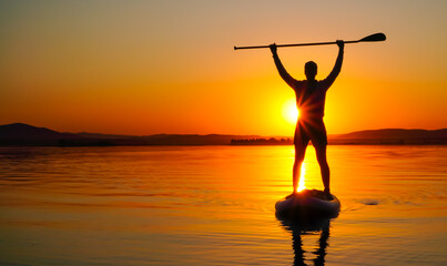 A young man is floating on a lake on a SUP board at sunset. The man's hands are raised up. Active recreation of a person in nature.