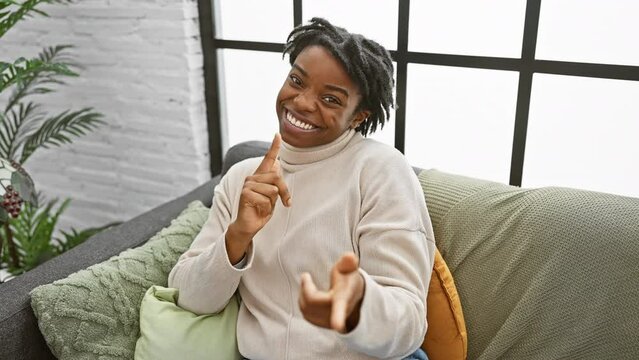 Young black woman radiates positivity sitting on home sofa. her dreadlocks, big smile, happy eyes and joyful gesture of pointing towards the camera for you create an infectious good vibe.