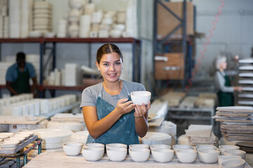 Portrait of positive young woman in apron working in ceramic workshop, holding crafted bowl,...