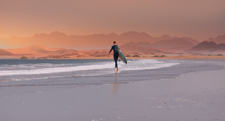 A male surfer walks on the beach with a surfboard in hand - Namib desert with Atlantic ocean meets near Skeleton coast - Namibia, South Africa - obrazy, fototapety, plakaty