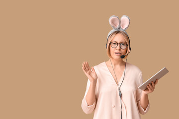 Surprised adult female operator in bunny ears with headset and tablet computer on beige background....