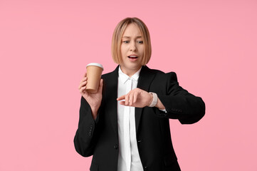 Worried adult woman with paper cup of coffee looking at wristwatch on pink background