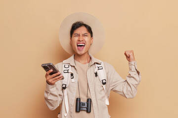A man in a hat smiles while holding a cell phone and binoculars