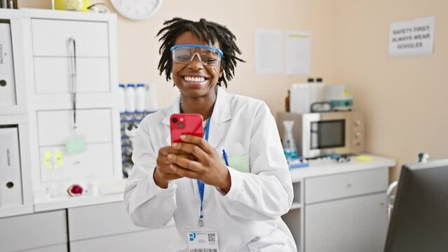 Cheery young black woman with dreadlocks flashing her toothy smile, ecstatic while working at the lab, expertly handling her phone with confident success.